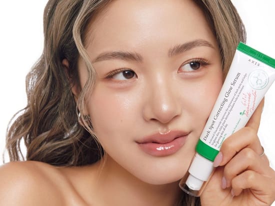 Her ses en kvinde, der holder AXIS-Y Dark Spot Correcting Glow Serum ved ansigtet, på en hvid baggrund.