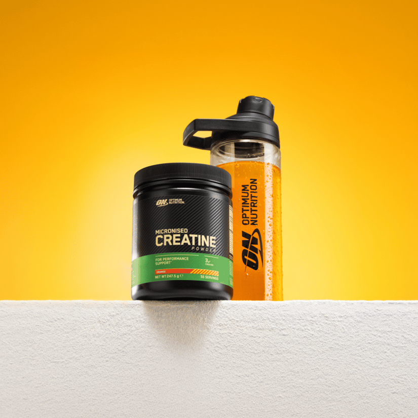 Bøtte med Optimum Nutrition Micronised Creatine og en shaker med orange drik står mod en kraftig orange baggrund.