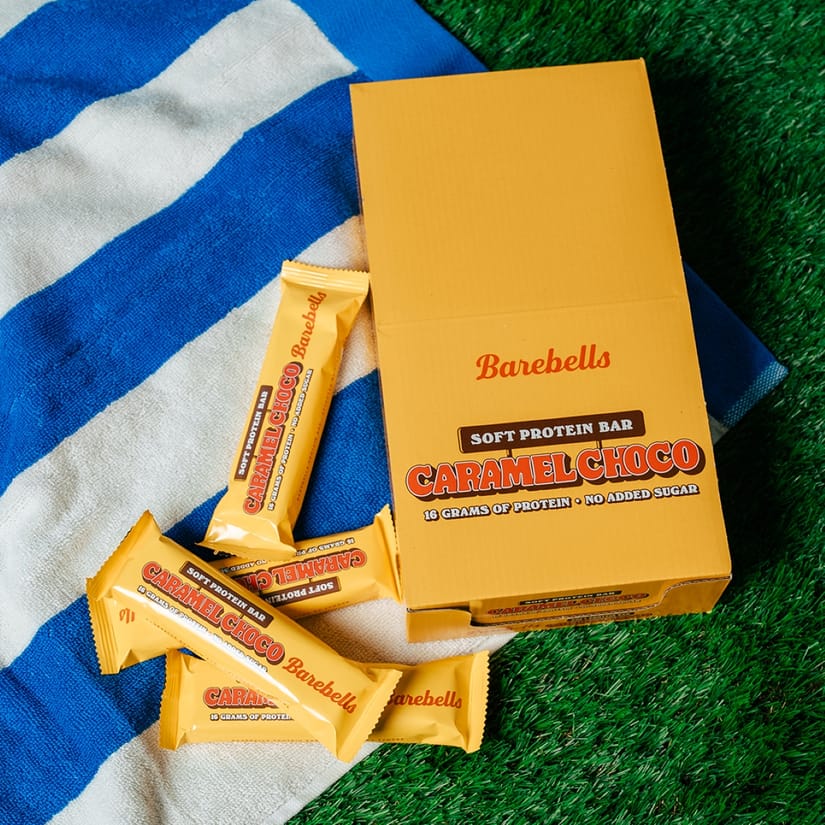 Barebells Caramel Choco-proteinbarer og en gul produktæske ligger på et blåt og hvidt håndklæde ved siden af grønt kunstgræs.