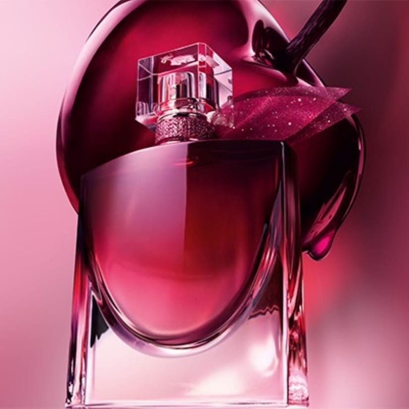 Her ses Lancôme La Vie Est Belle Vanille Nude Very Cherry i en rosa parfumeflakon med transparent låg og en mørkerosa, glitrende sløjfe. Bag flakonen ses et stort mørkerødt kirsebær mod en rosa baggrund.